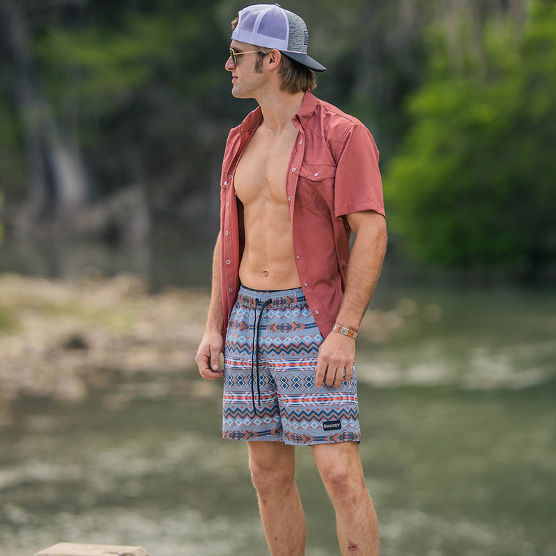 male model wearing the Bigwake grey, red, blue Aztec pattern board shorts, sienna sol shirt, and grey Hooey cap at the river
