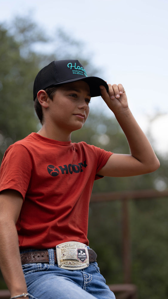 youth model posing in hooey red tee with charcoal and teal hat