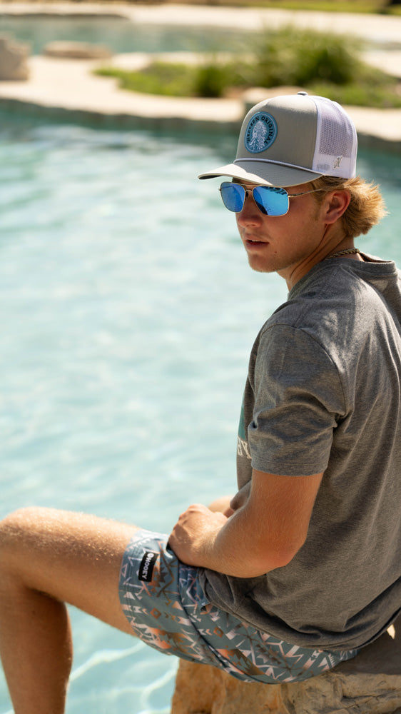 male model wearing Hooey grey, blue, white multi pattern board shorts, grey tee, and grey and blue hat seated on edge of pool with foot in water