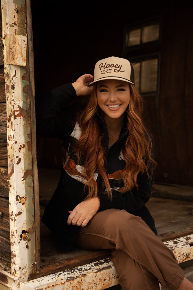 female model wearing black jacket with desert scene and tan and brown Hooey hat