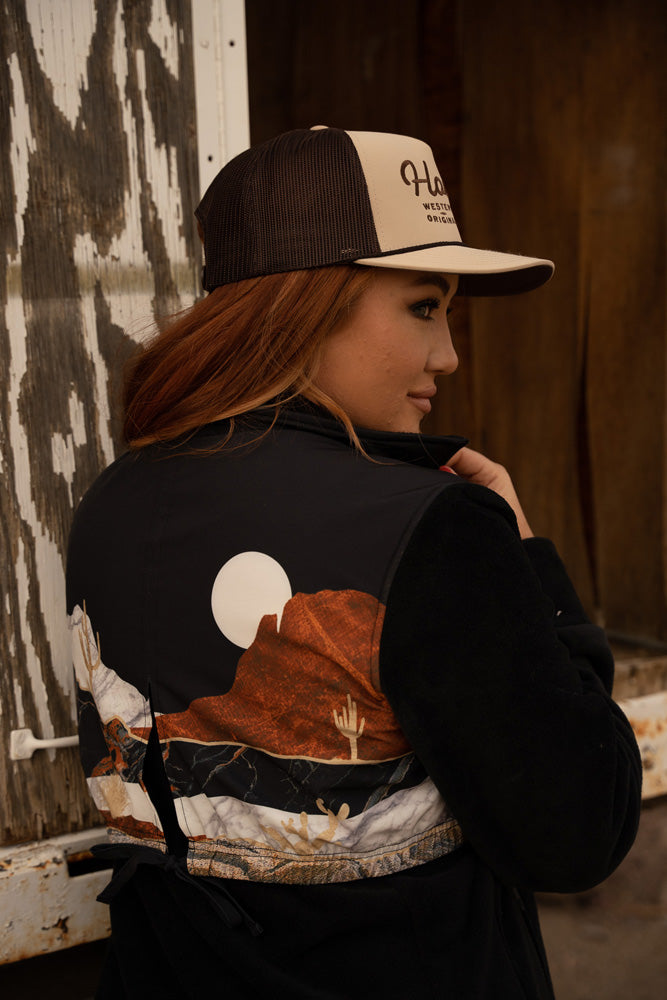 female model wearing black jacket with desert scene on back and tan and brown Hooey hat