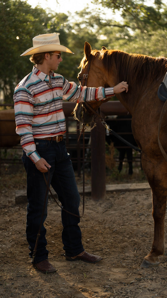 male model earing dark wash jeans, cowboy boots and hat, with white, red, blue striped sol shirt. posing with a horse in an outdoor arena 
