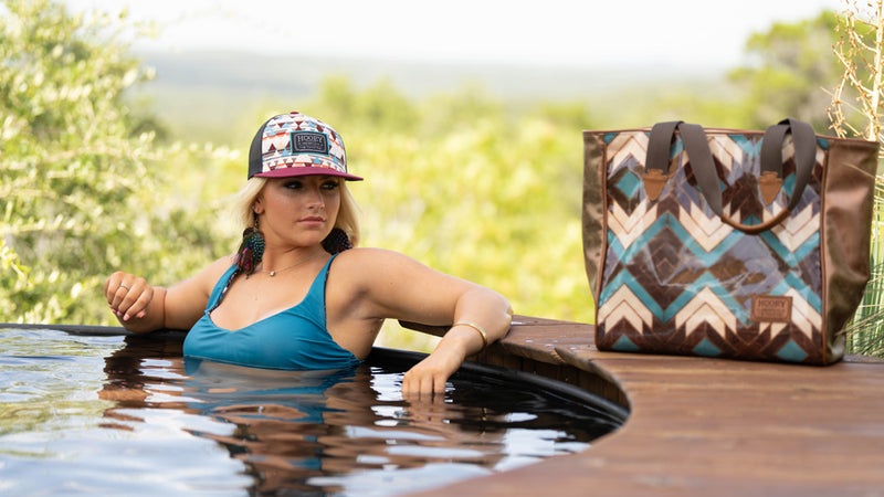 female model posing in pool wearing reversible, blue swim suit, RLAG hat with aztec pattern tote bag