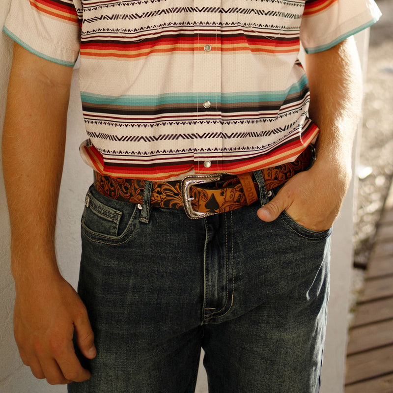 close up of Hooey's tooled leather belt, medium washed jeans, and multi colored sol shirt