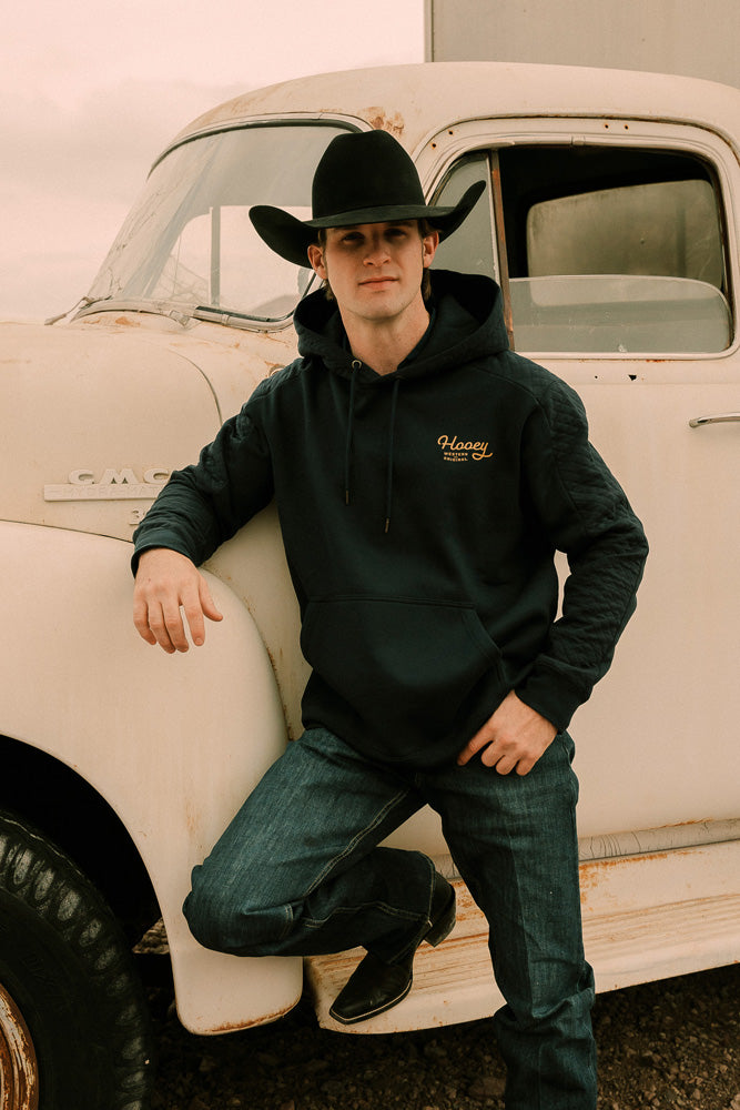 model wearing the canyon navy with gold hoody leaning against an old white truck