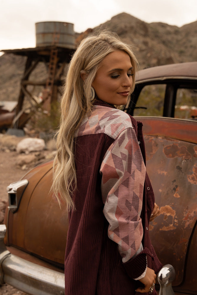 blonde female model looking over her shoulder wearing burgundy corduroy jacket with orange and blue Aztec pattern sleeves in front of rusty old junk car