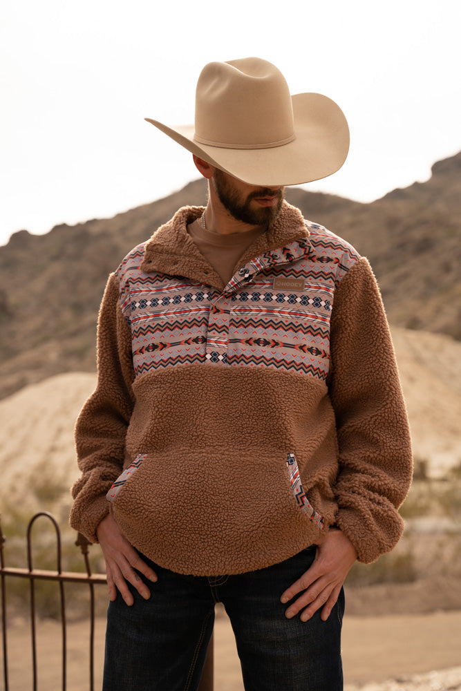 Male model wearing black jeans, straw cowboy hat, and the Hooey Sherpa Fleece Pullover in tan with blue, red, white, Aztec pattern on collar, chest, and pocket area in an outdoor mountainous setting