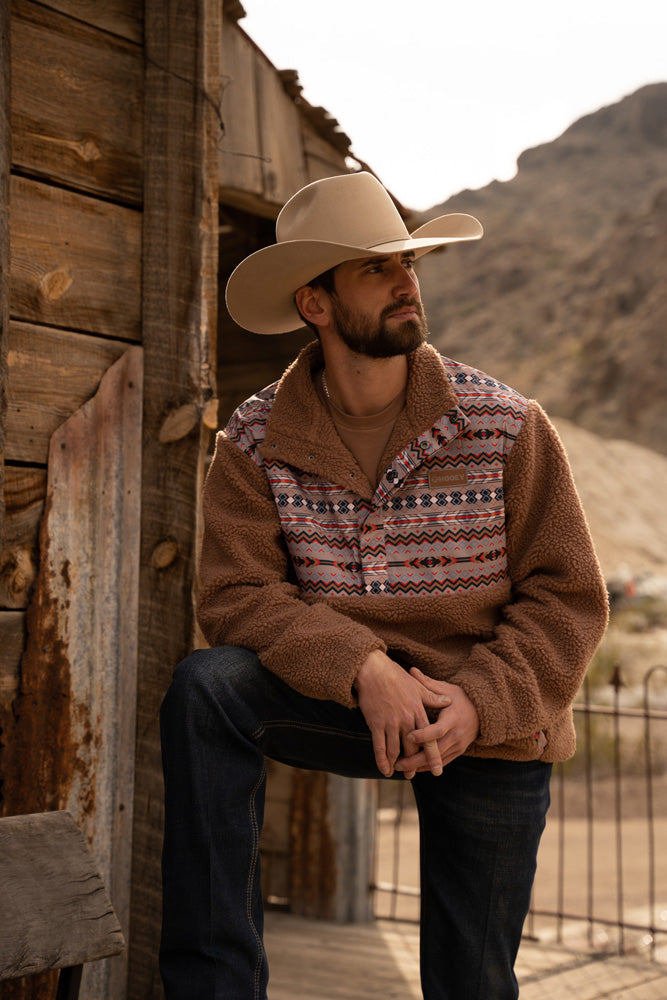 male model wearing a straw cowboy hat, dark wash jeans, and Hooey Sherpa Fleece Pullover in tan with blue, red, white, Aztec pattern on collar, chest, and pocket area with foot propped up against a rustic wood building