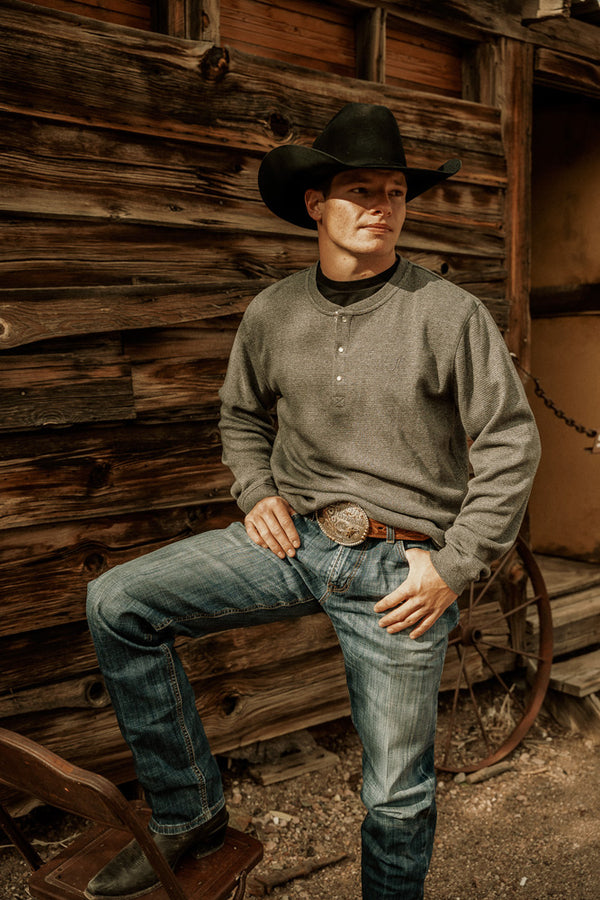male model wearing the Asphalt long sleeve thermal Henley shirt, jeans, black cowboy boots and matching cowboy hat staged in front of a rustic log cabin