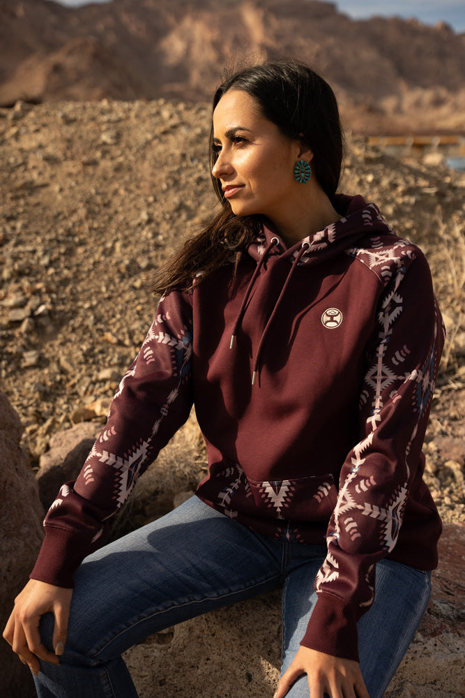 female model wearing the Summit maroon hoody with cream and blue aztec pattern on pocket and sleeves and hood with jeans seated on a bolder