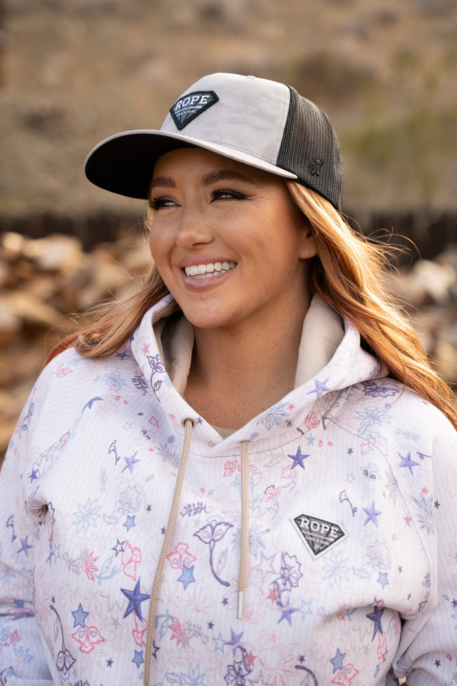 Rope Like A Girl white hoody with star and floral pattern and grey and black RLAG hat worn by a brunet, female model in outdoor setting with blurred background
