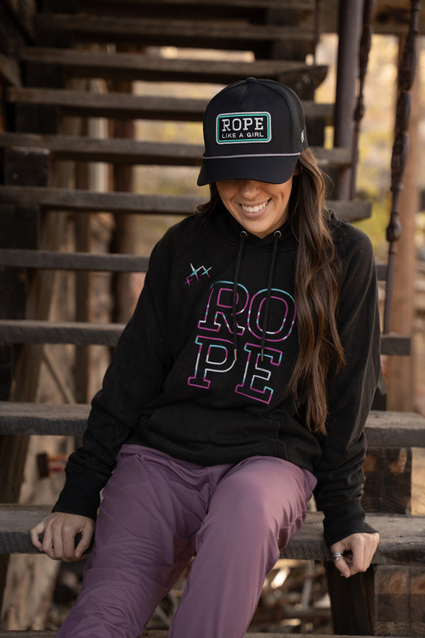 lifestyle image of a burnet female model wearing the Rope Like A Girl black hoody with multi colored logo, purple pants, and black Rope Like A Girl hat with teal stitching, sitting on an outdoor starcase