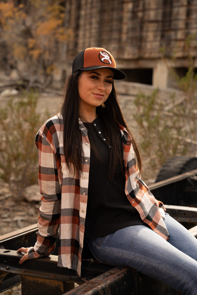 female model wearing orange and black hat with black and white hooey logo, orange, cream, and grey plaid shacket, black henley and blue jeans seated on a trailer chasey