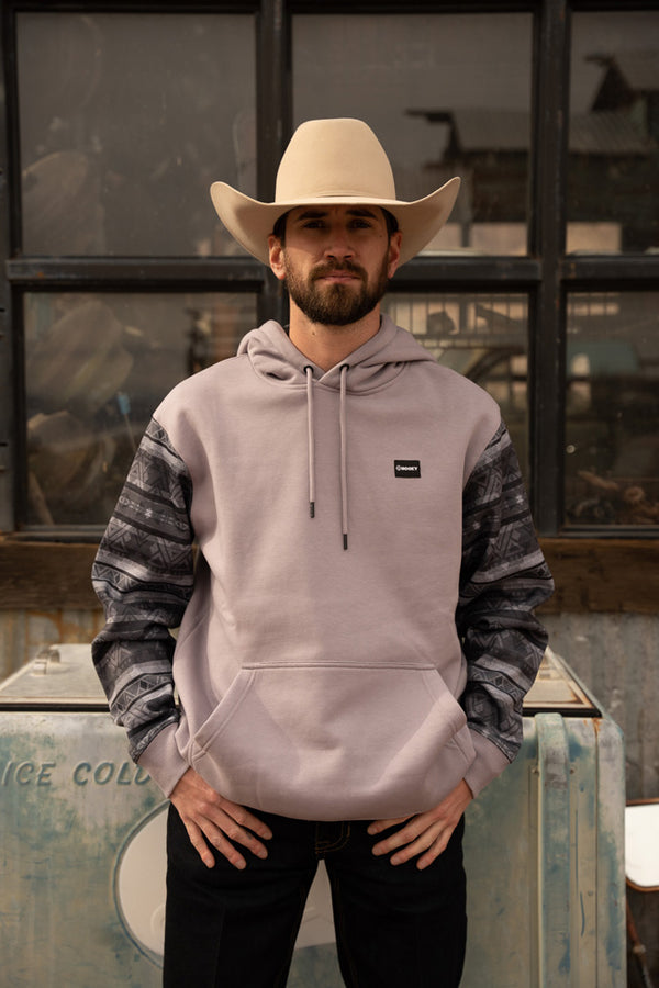 male model wearing a cream felt hat, black jeans, and summit grey hoody with grey and black aztec pattern on sleeves and hood infront of a multi pane window