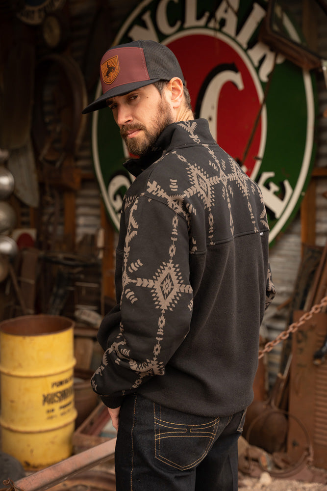 back of male model wearing the Stevie black pullover with black and grey Aztec pattern on collar and sleeves in rusting looking garage setting