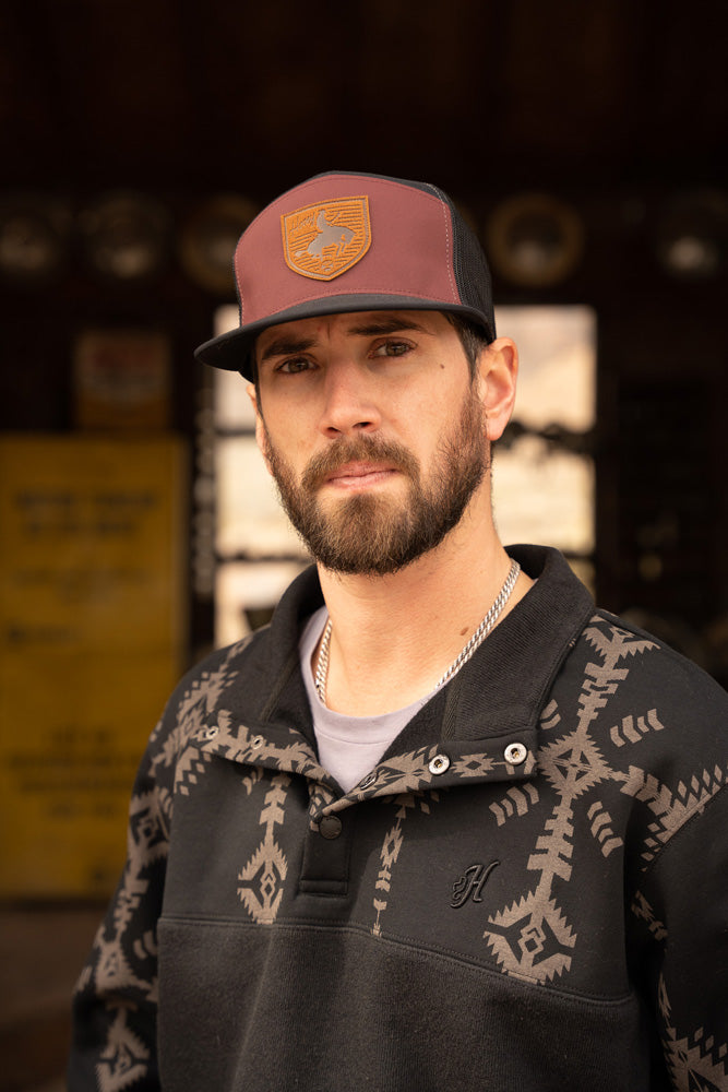 male model wearing the Stevie black pullover with black and grey Aztec pattern on collar and sleeves and maroon/black hat with leather patch in rustic building