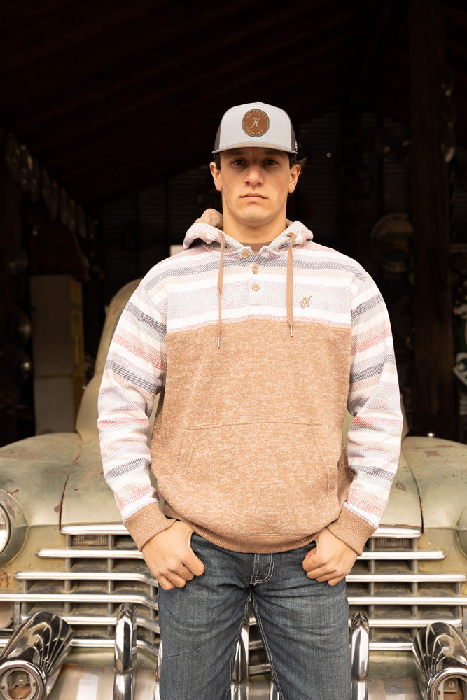 male model wearing jeans, white and grey hat with leather patch and Jimmy tan hoody with pink, blue, navy, teal, tan, cream serape pattern on collar and sleeves posed in front of an antique car in rustic garage