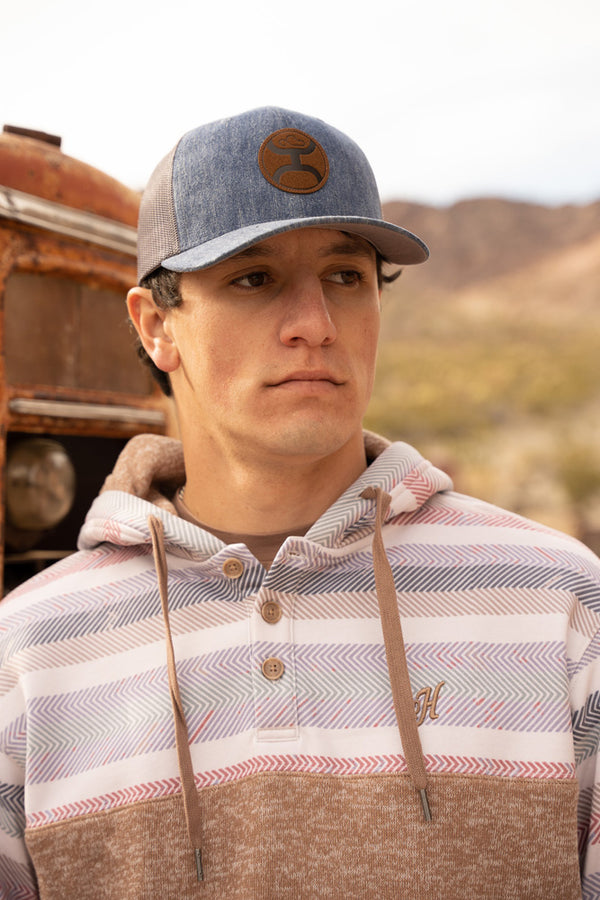 male model wearing denim hat with leather hooey patch and Jimmy tan hoody with pink, blue, navy, teal, tan, cream serape pattern on collar and sleeves in outdoor setting