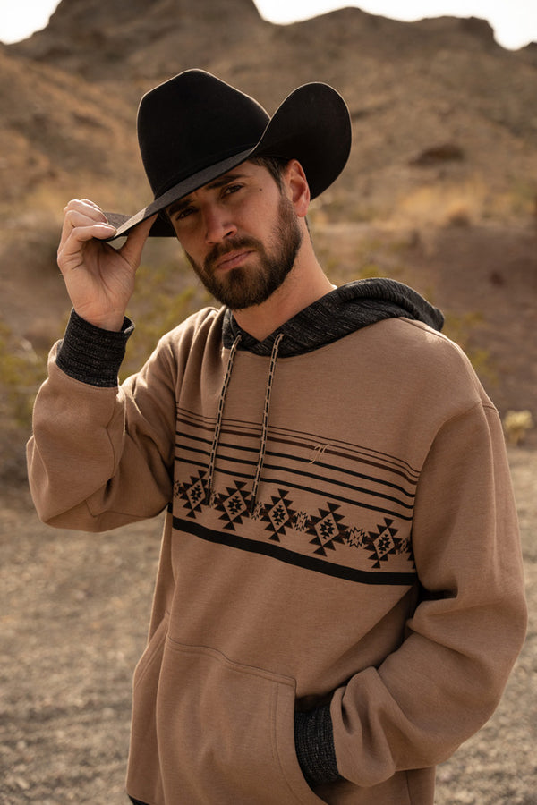 male model wearing the Taos tan hoody with brown aztec pattern across chest and heather black cuffs and hood with black felt cowboy hat in desert setting