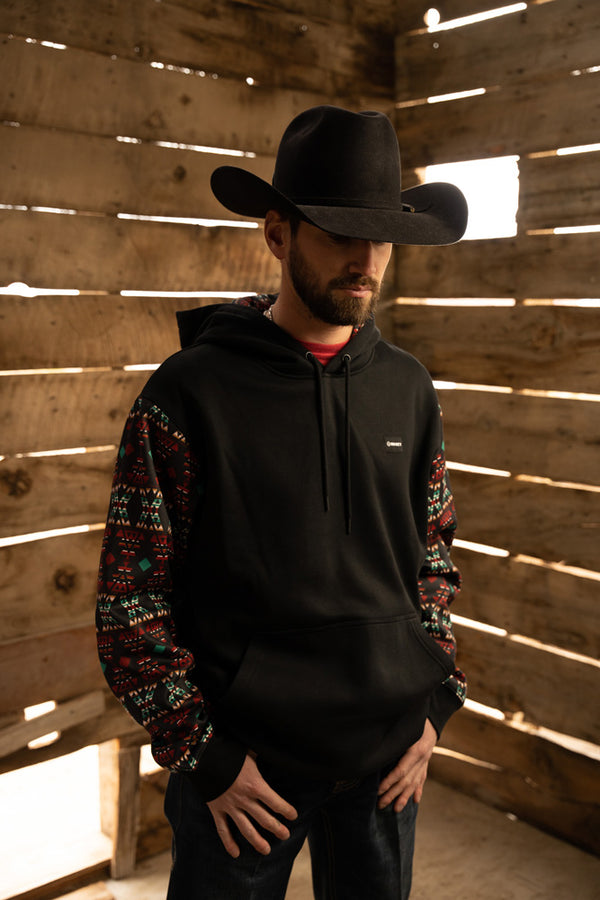 male model wearing the summit black hoody with red and blue aztec pattern on sleeves and black felt cowboy hat in rustic wood structure