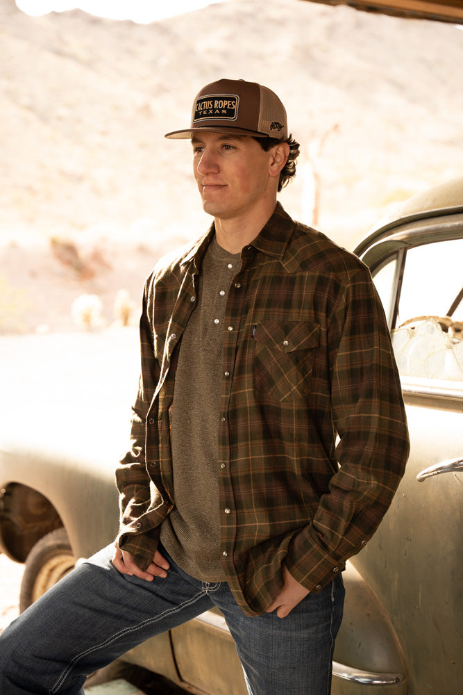 male model wearing dark wash jeans, olive henley and green/brown plaid shacket and brown and tan cactus ropes cap
