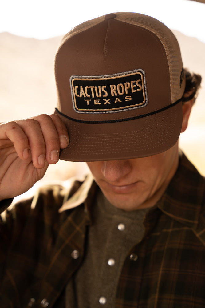 male model wearing brown plaid shakcet with olive henley and brown/tan cactus ropes hat pulled over eyes with right hand