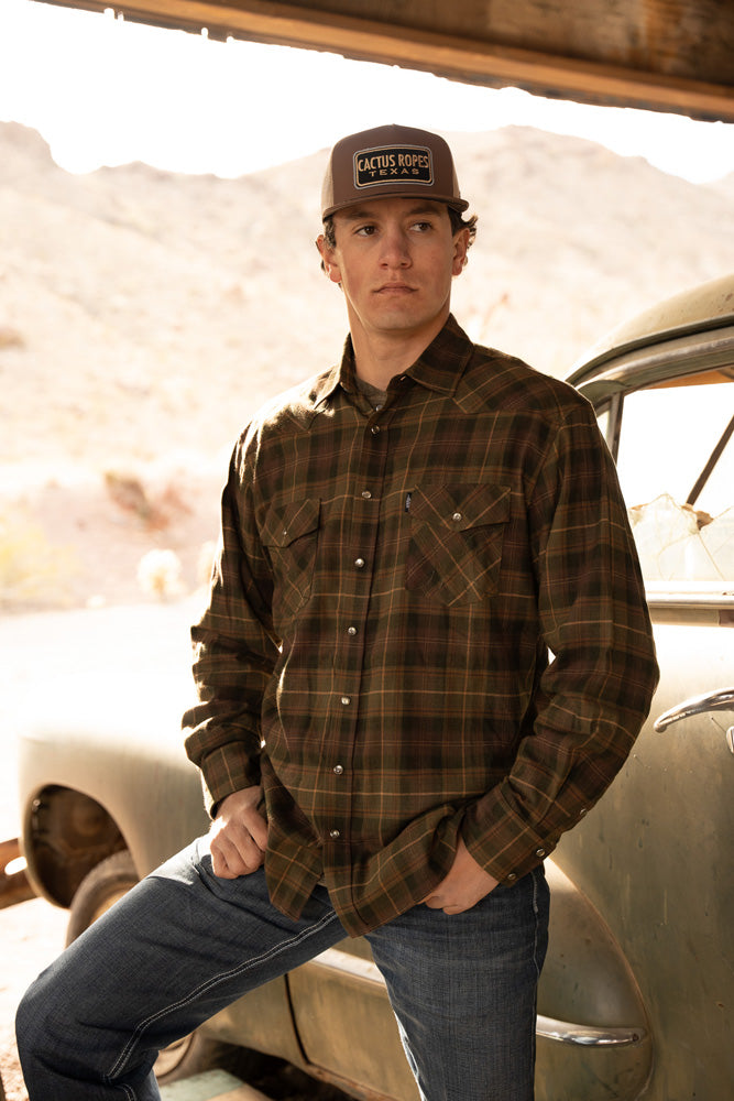 male model wearing dark wash jeans, olive henley and green/brown plaid shacket and brown and tan cactus ropes cap