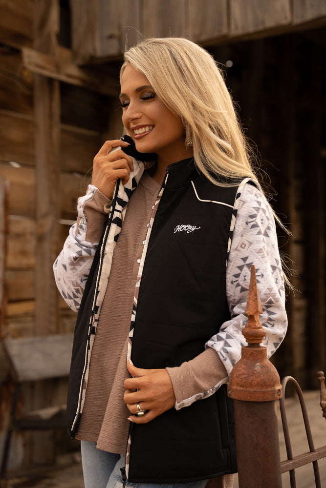 female model showing off her black Hooey vest with black and white aztec print lining, tan and white henley, and light washed jeans in rustic setting