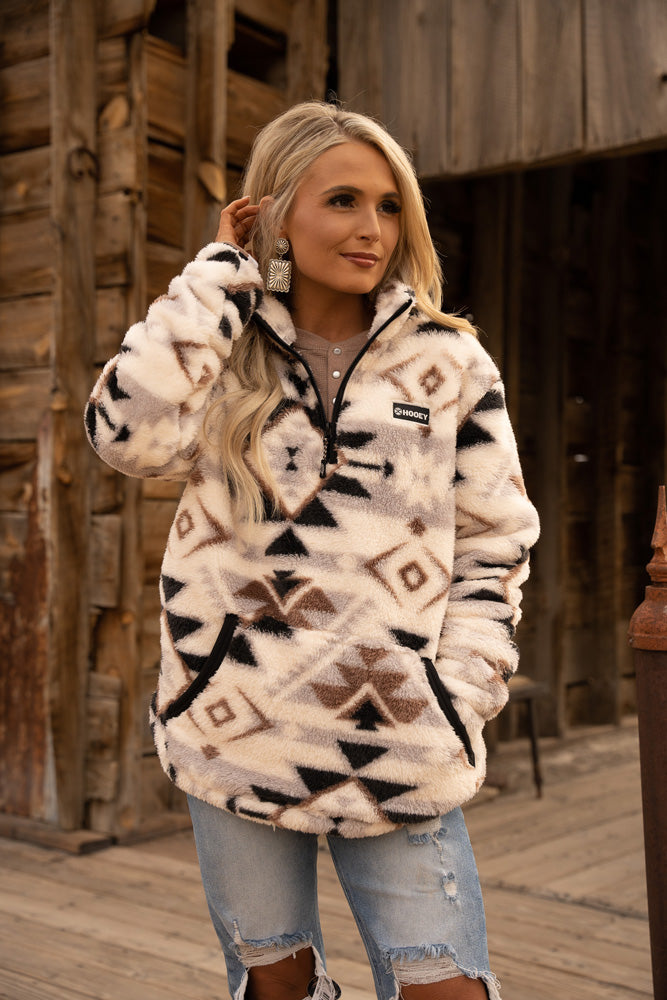 female model posing in ripped jeans with cream, grey, brown, black Aztec pattern fleece pullover in rustic setting