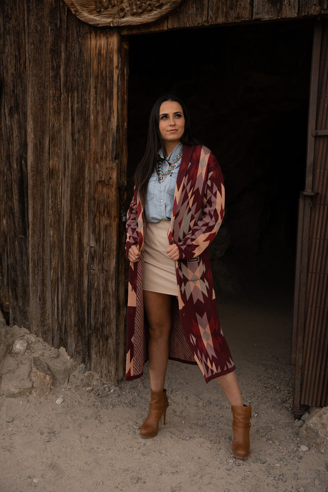 female model posing in doorway of rustic wood structure sporting burgundy aztec duster