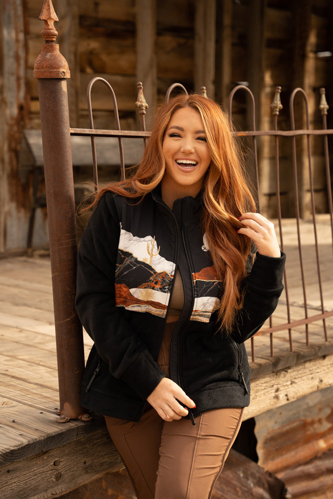 female hooey model wearing brown pants and desert scene zip jacket posed in front of wood porch 