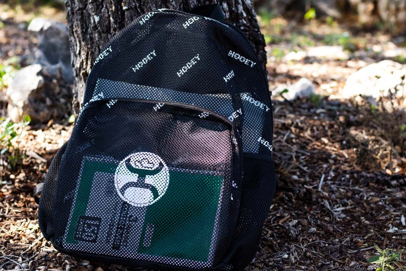 Nitro Mesh backpack in black with white Hooey logos all over and large Hooey emblem on front pocket leaned agains a treen with a notebook inside the backpack