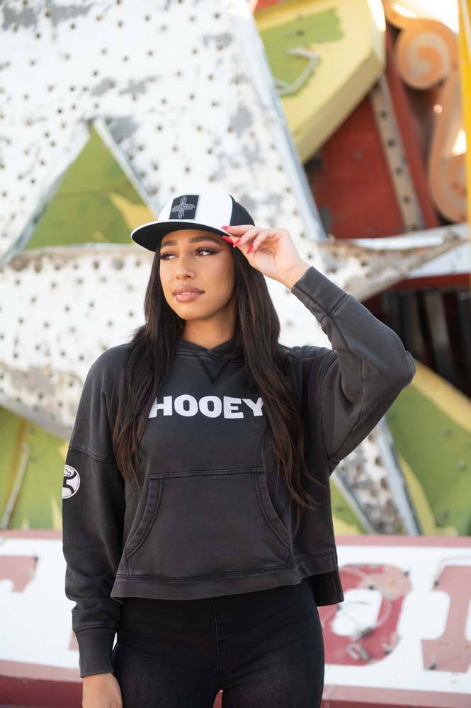 female model wearing the Roomy black hoody with white logo, black and white hat, and black pants posed infront of signs