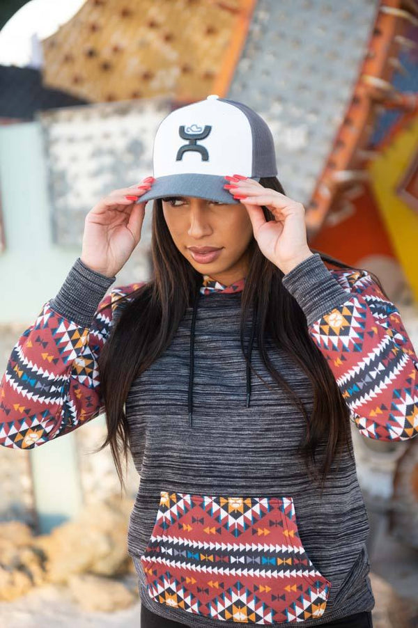 female model wearing the Summit heather charcoal with red, tan. and blue aztec patter on sleeves, pocket, and hood with white and grey hat