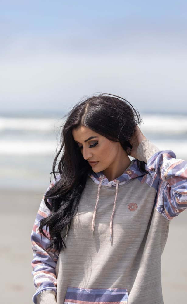 female model wearing the summit cream hoody with pink, grey, orange aztec pattern on pocket, sleeves, and hood in beach setting