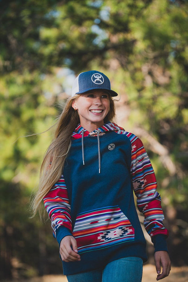 female model wearing the Taos heather navy hoody with red, orange, white, blue purple aztec and serape pattern on sleeves and hood with jeans and denim hooey hat in out door setting