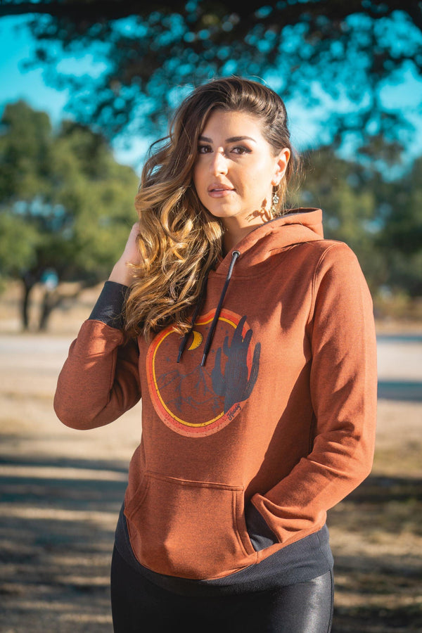 female model wearing the Marfa heather orange hoody with orange, yellow, black artwork, cuffs, and hood lining, black leather pants, in outdoor setting