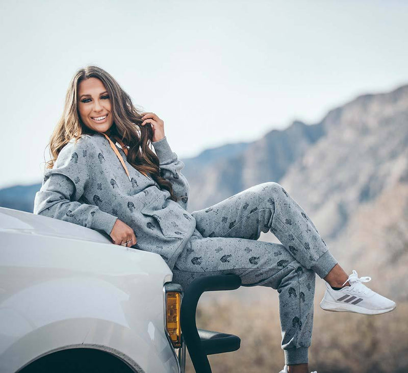 female model wearing Plains joggers in grey with black pattern and matching top posed on hood of white truck