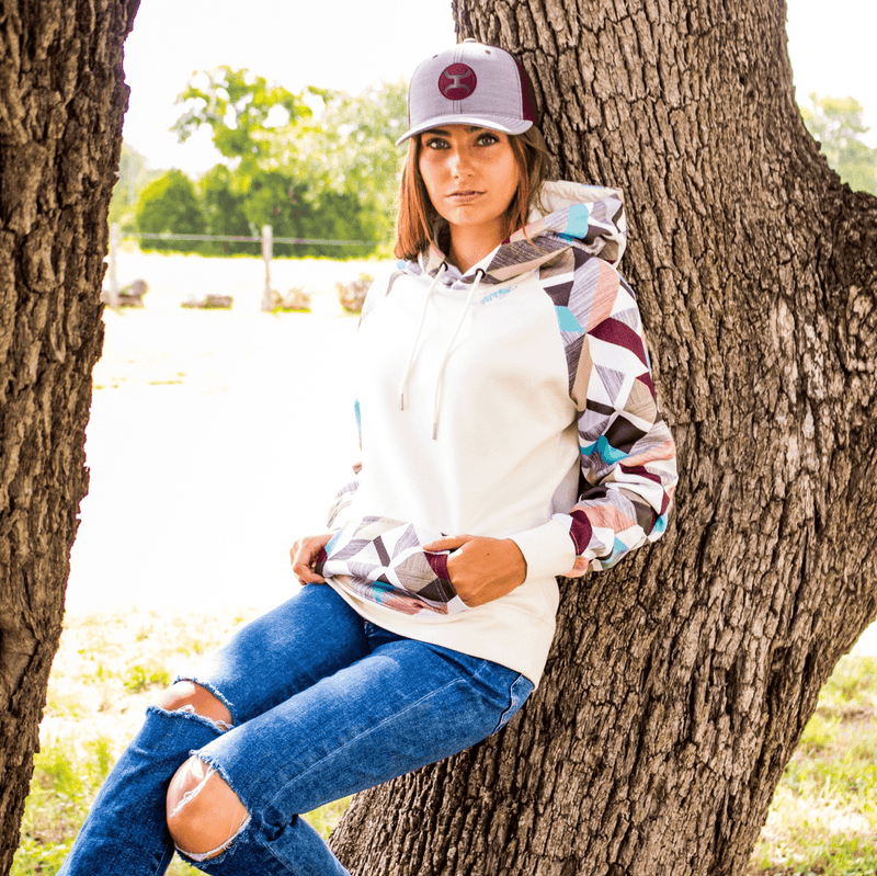 female model wearing the Morocco white hoody with tan, orange, blue, grey, purple patten on sleeves, pocket, and hood with jeans and grey/maroon hooey hat leaning against a tree