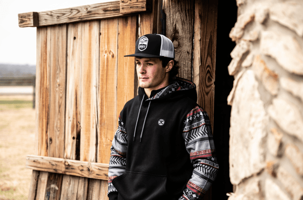 Cole Reiner stands outside a rustic wooden building wearing a black vest and black baseball hat.