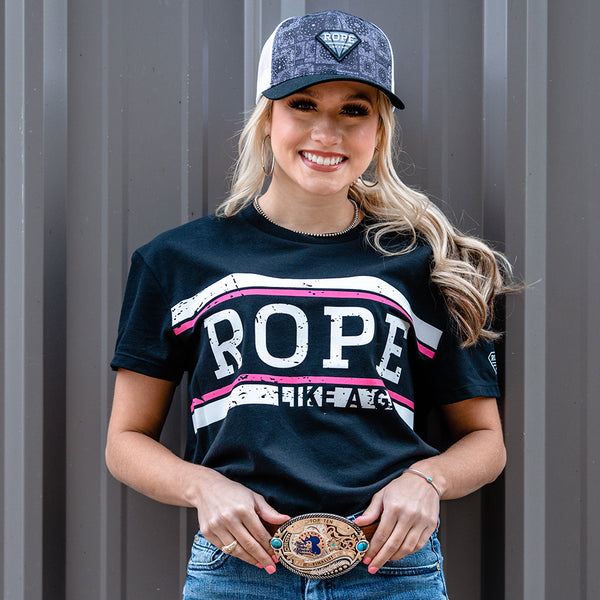 lifestyle image of a blond female model leaned against a metal building sporting the Rope Like A Gril black tee with white and pink logo across the chest, blue jeans, gold buckle, grey and white multi pattern RLAG hat