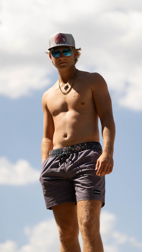 male model posing in Hooey hat and grey board shorts with hand in pocket and blue sky in the background