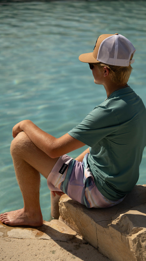 male model wearing pink/purple/blue serape board shorts with green tee and tan and white hooey hat seated on edge of pool with right foot in water. facing away from the camera
