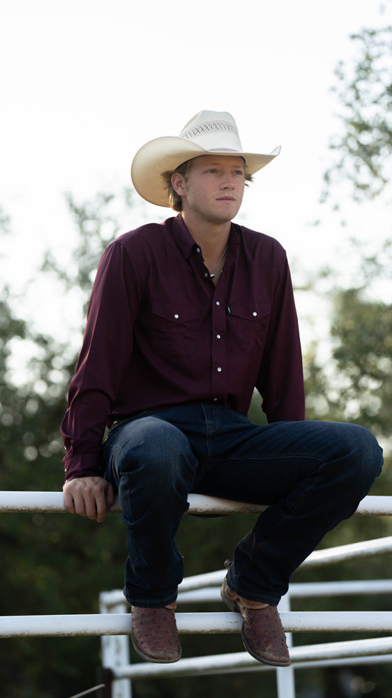male model wearing cowboy boots with straw cowboy boots and maroon sol shirt with dark wash jeans seated on white pipe fence