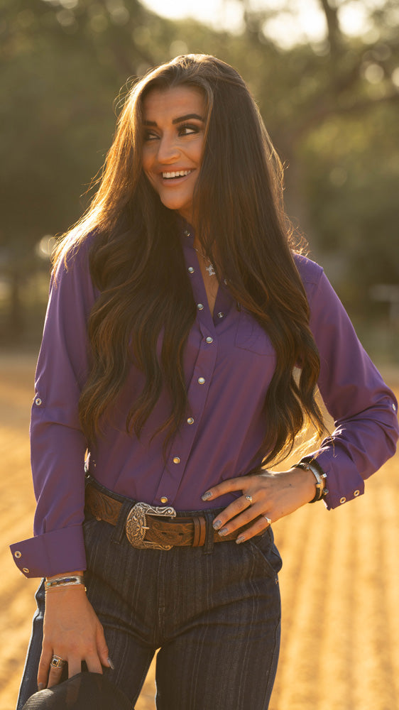 close up of female model wearing purple sol shirt with hooey trousers and brown belt, posing in outdoor rodeo arena
