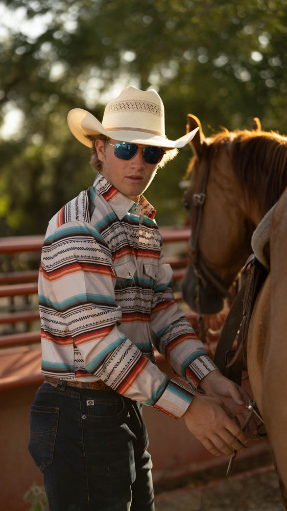 male model wearing Hooey's cream with serape pattern sol shirt, dark wash jeans, brown leather belt, and straw cowboy hat, and posing with a horse in  an outdoor setting