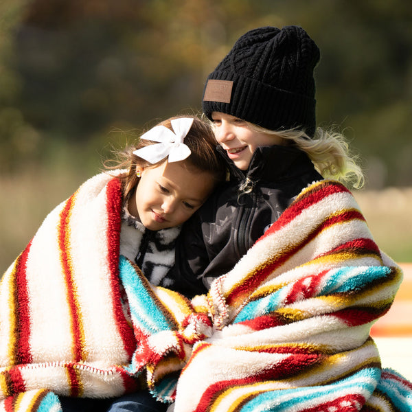 Two young girls wrapped in the new Hooey fleece blanket in serape print posing in an outdoor setting