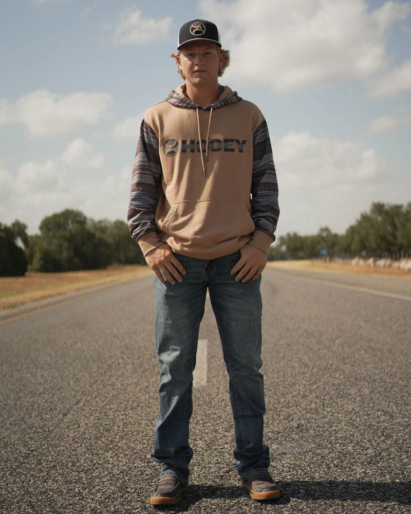young male model wearing Hooey loopers, medium wash jeans, tan/blue/red hooey hoody, with black/gold/white hat posting on paved road
