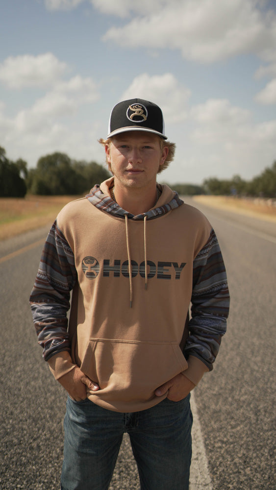 male model wearing black and white hooey hat with tan and grey Hooey hoody and medium wash jeans posing on paved road