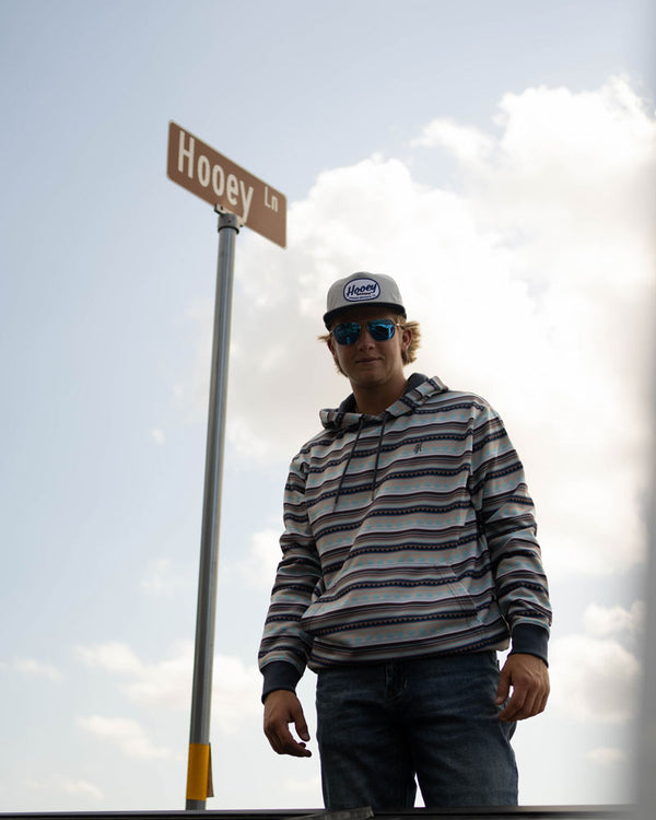male model wearing blue jeans with red/grey/white Hooey striped hoody and grey and navy hooey hat posed next to Hooey lane road sign